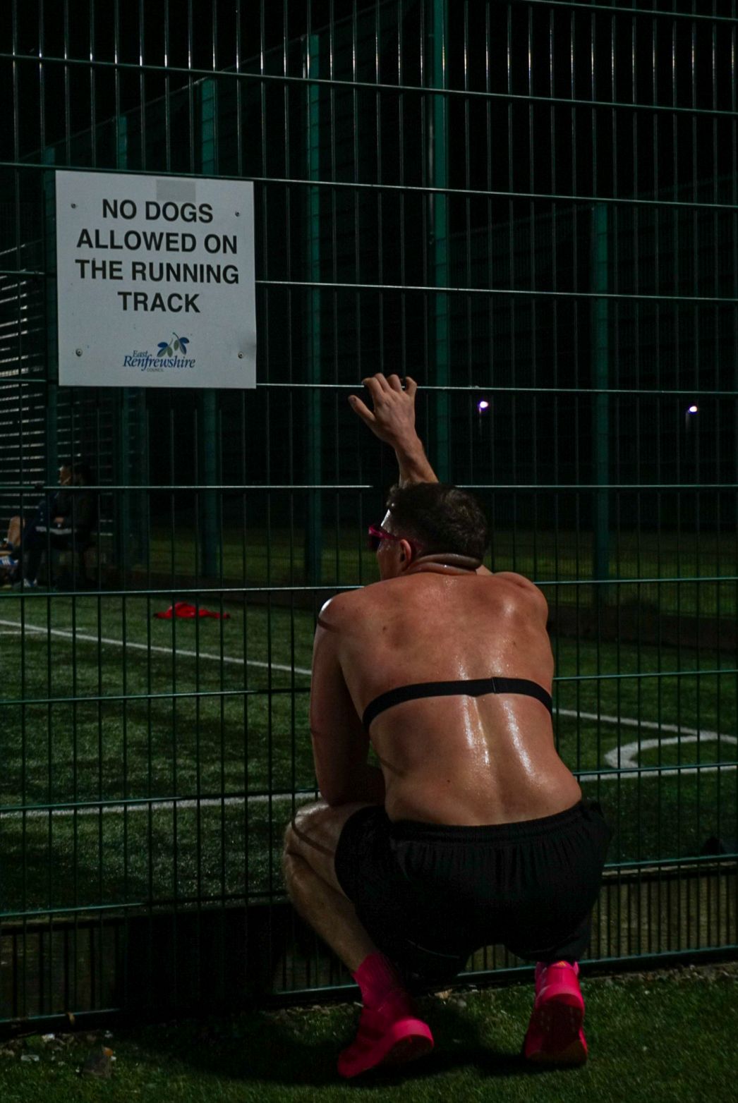 Shirtless male runner resting against a fence beside a no dogs allowed on running track sign.