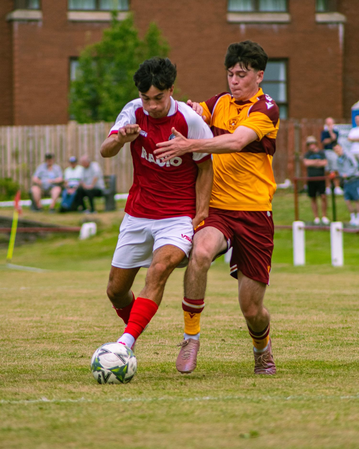 Two soccer players battle for the ball during a competitive match, one in red and white, the other in gold and maroon.