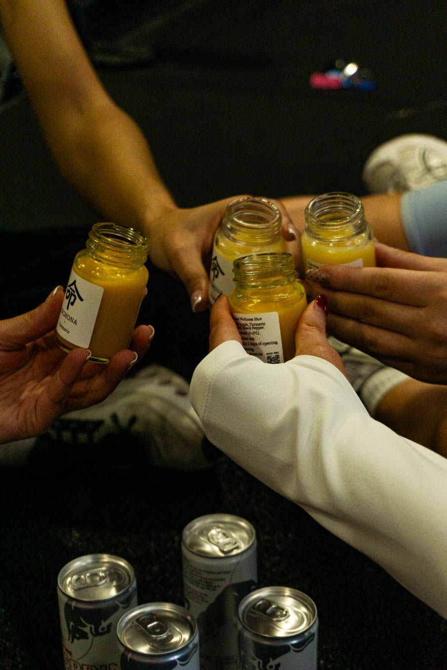 People toasting with small jars of yellow smoothie drinks over canned beverages on a dark surface.