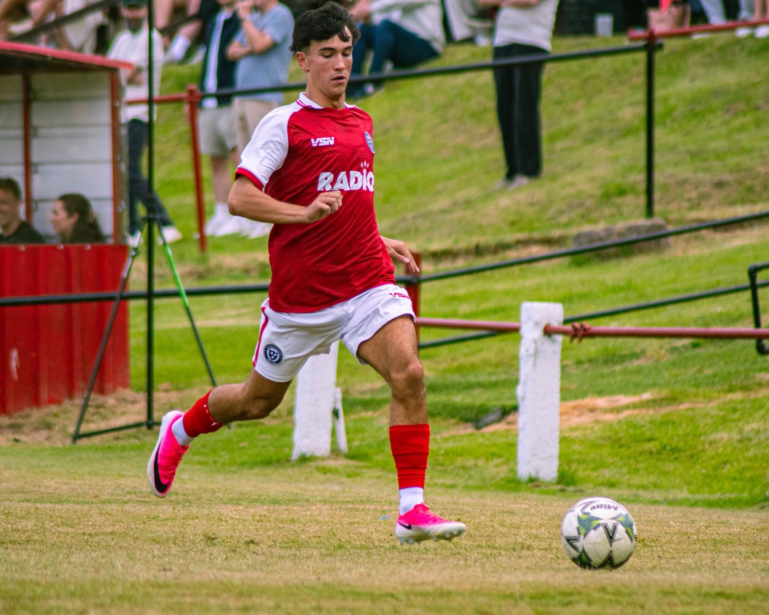 A young soccer player in a red and white uniform dribbles a ball on a grass field during a match.