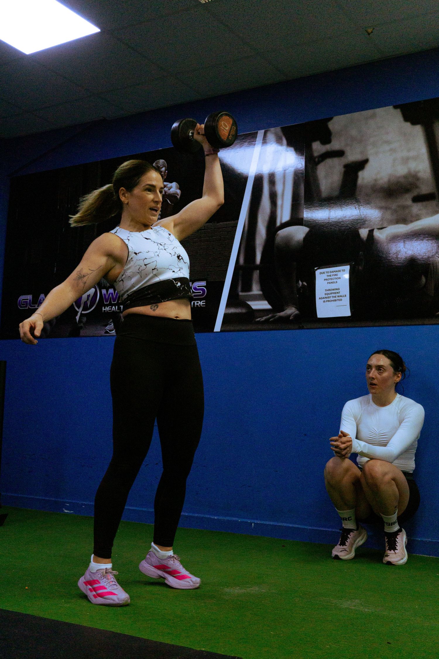 Woman lifting dumbbell overhead in gym while trainer observes during fitness workout session.
