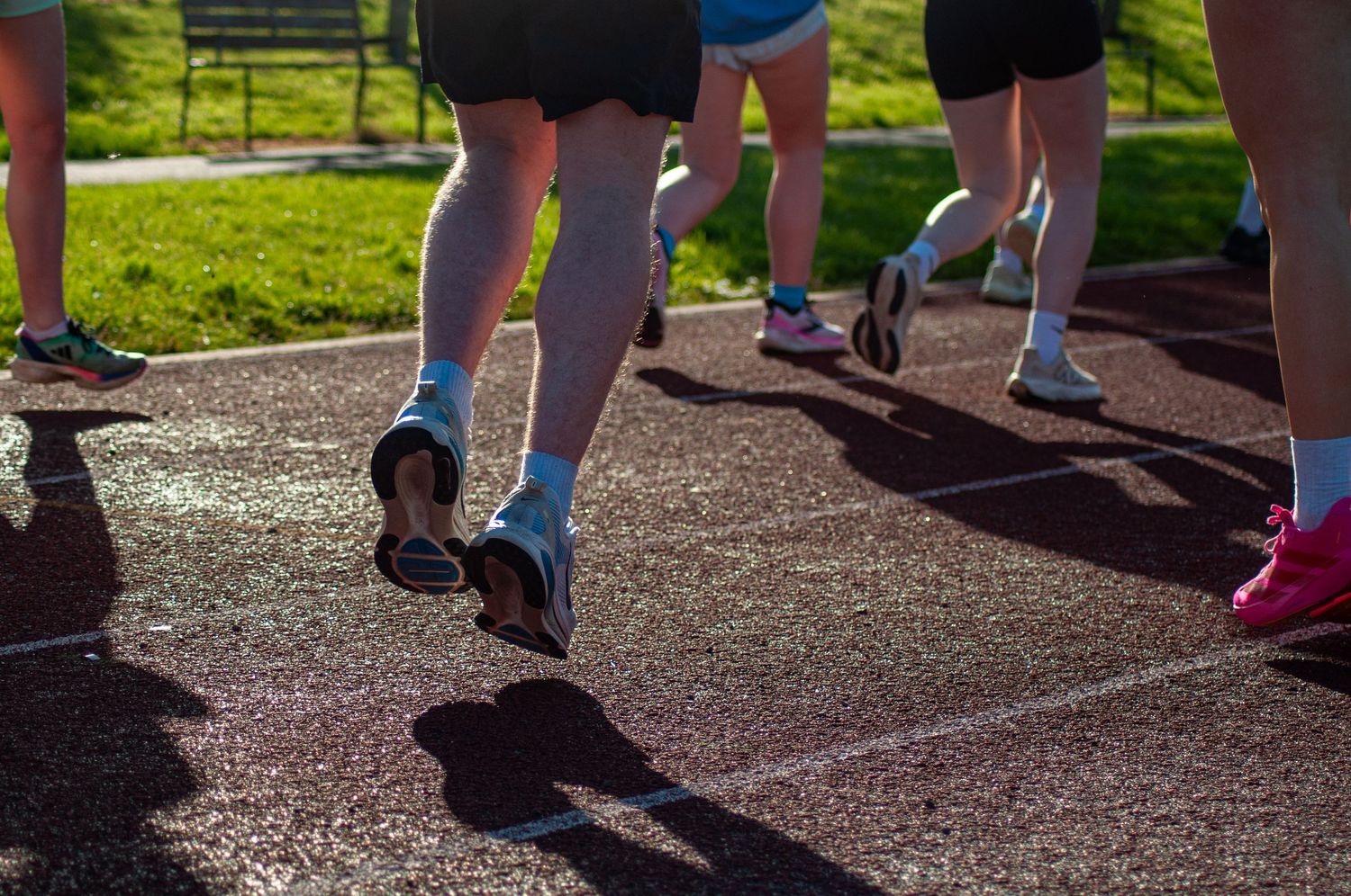 Close-up of runners feet and legs on a red athletics track casting long shadows.