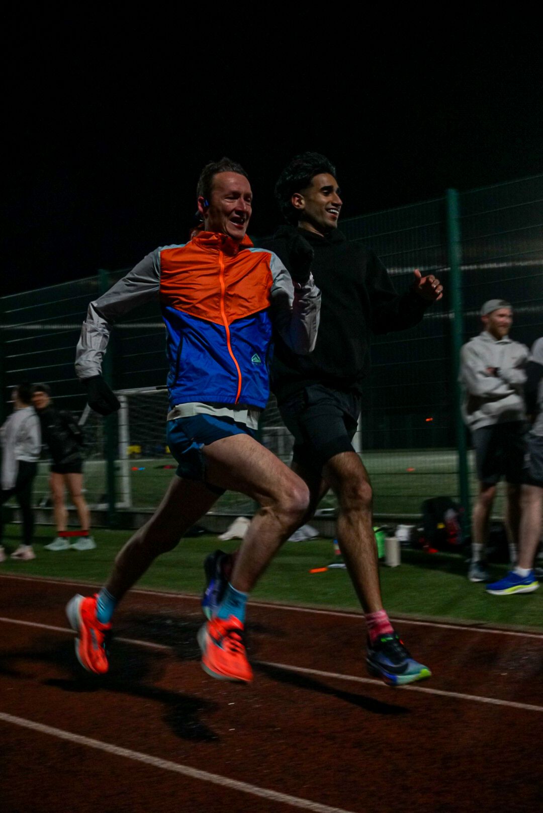 Two male runners sprint together on a floodlit athletics track at night, wearing colorful running shoes.