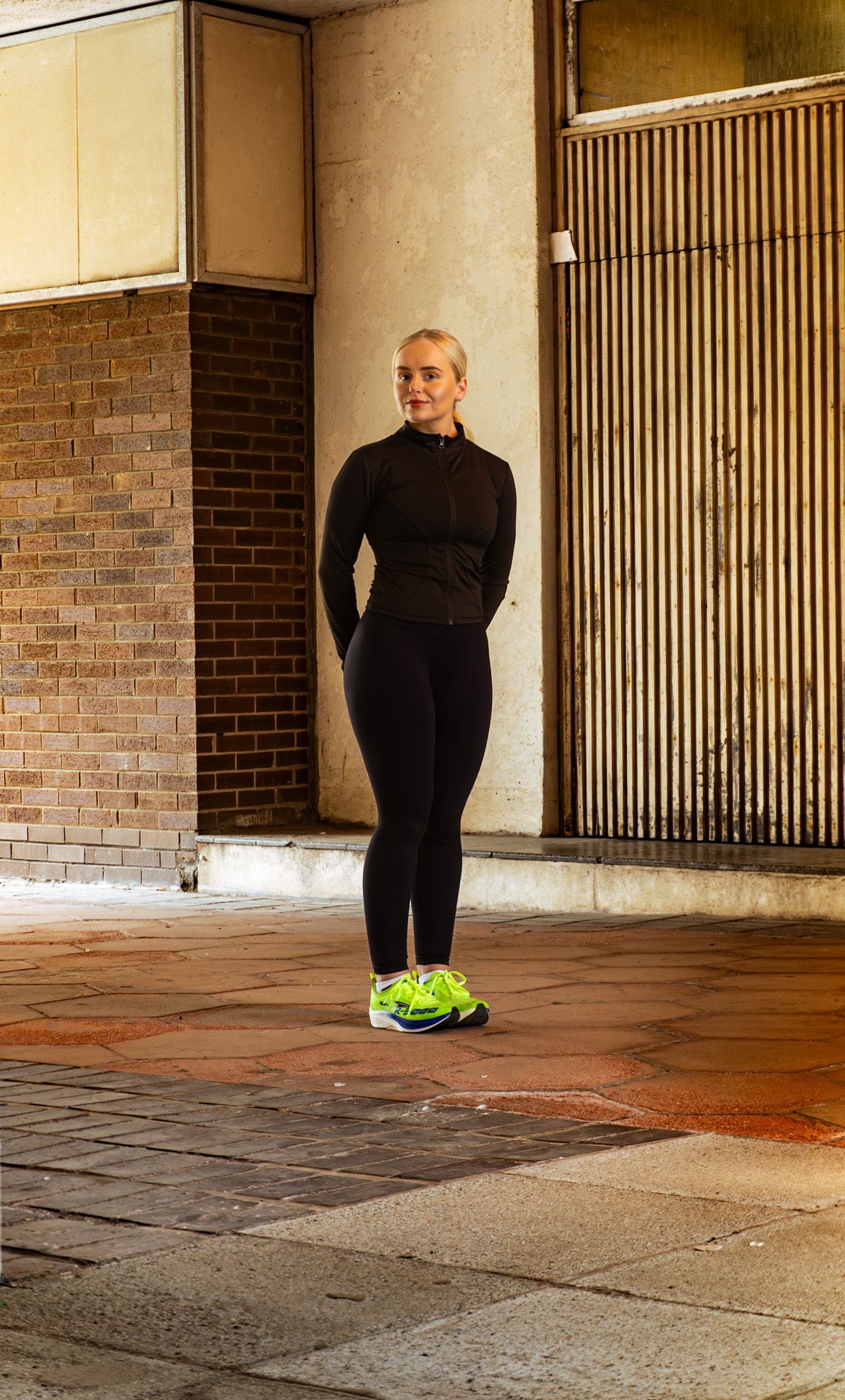 Blonde woman in black athletic leggings and jacket standing in urban setting with neon green shoes.