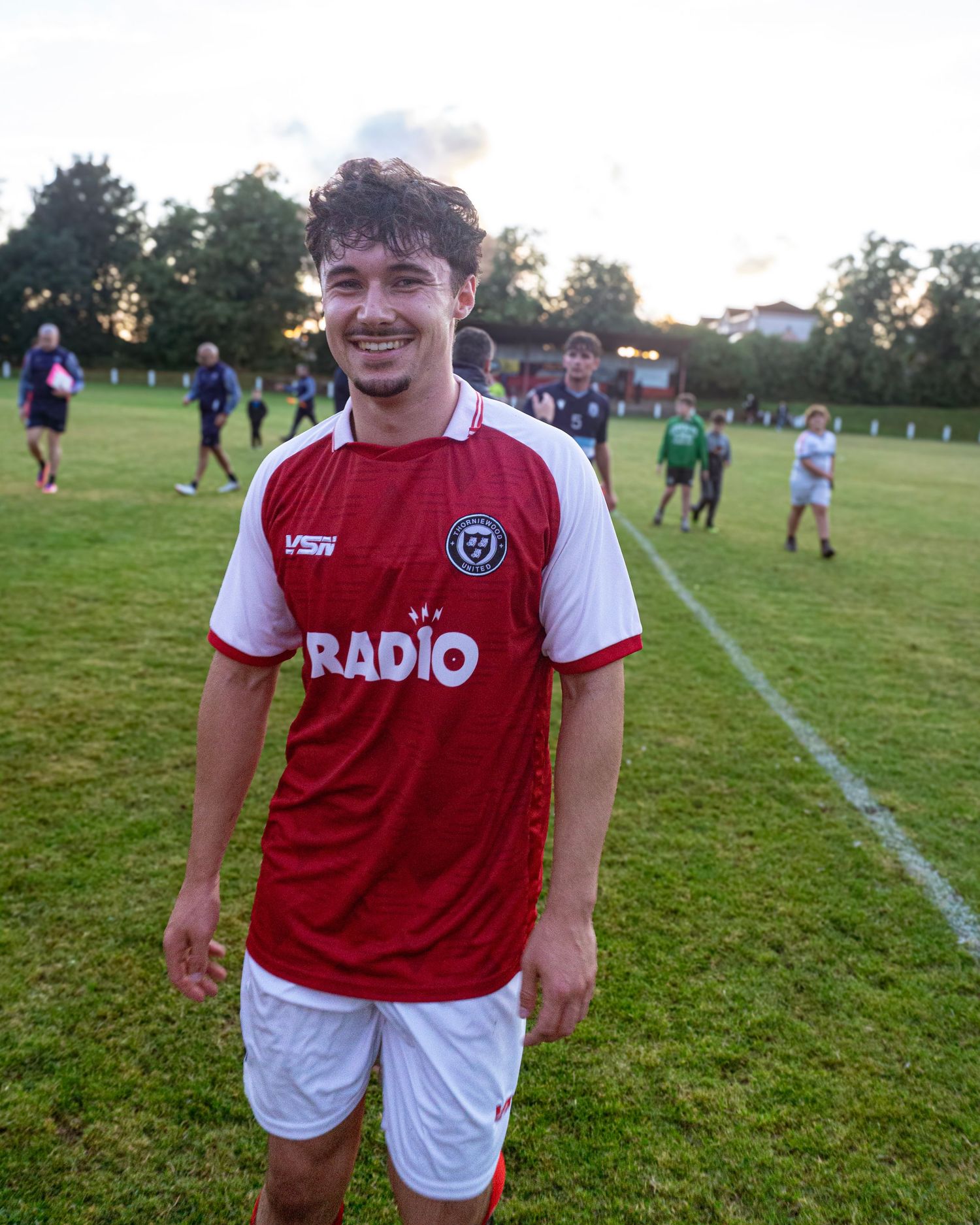 Young male soccer player smiles in red and white RADIO jersey on a football pitch at dusk.
