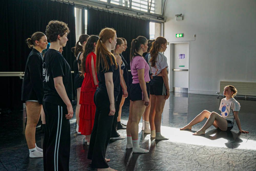 Blurred motion of dancers moving energetically past chairs and mirrors in a bright dance rehearsal studio.