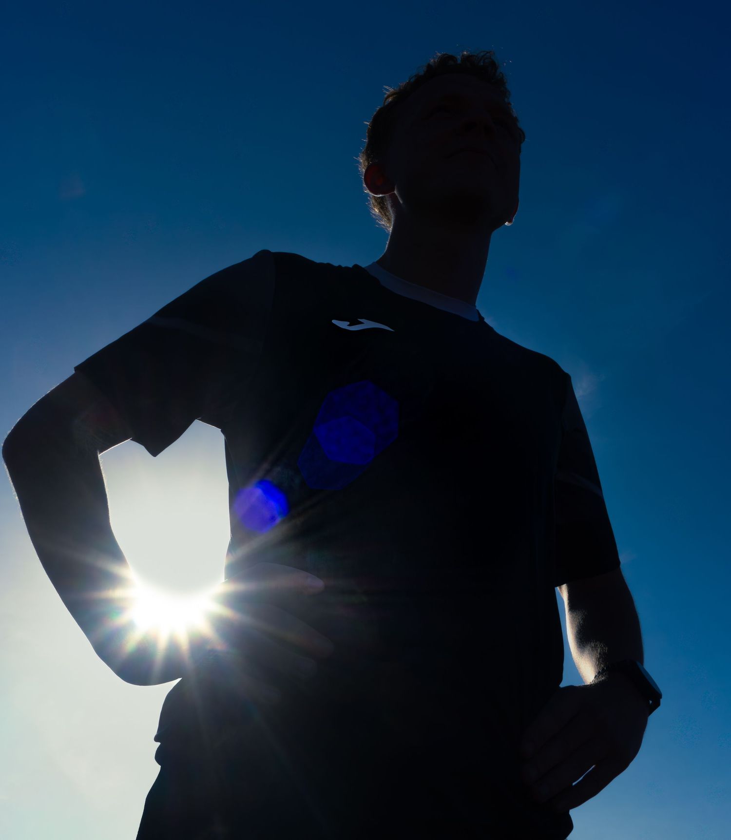 Silhouette of a male athlete in a Joma shirt backlit by bright sunlight against a deep blue sky.