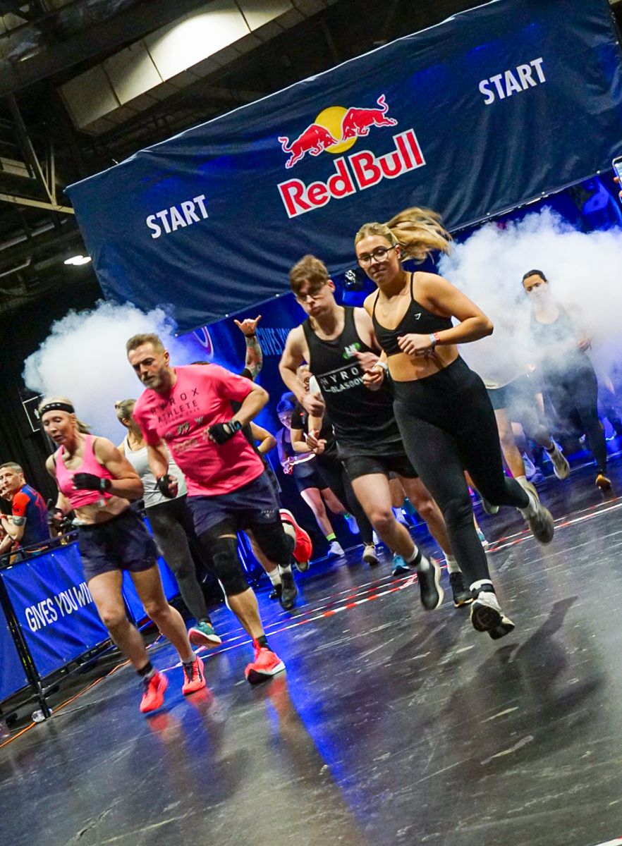 Athletes burst from the start line under a Red Bull banner at a HYROX fitness competition.
