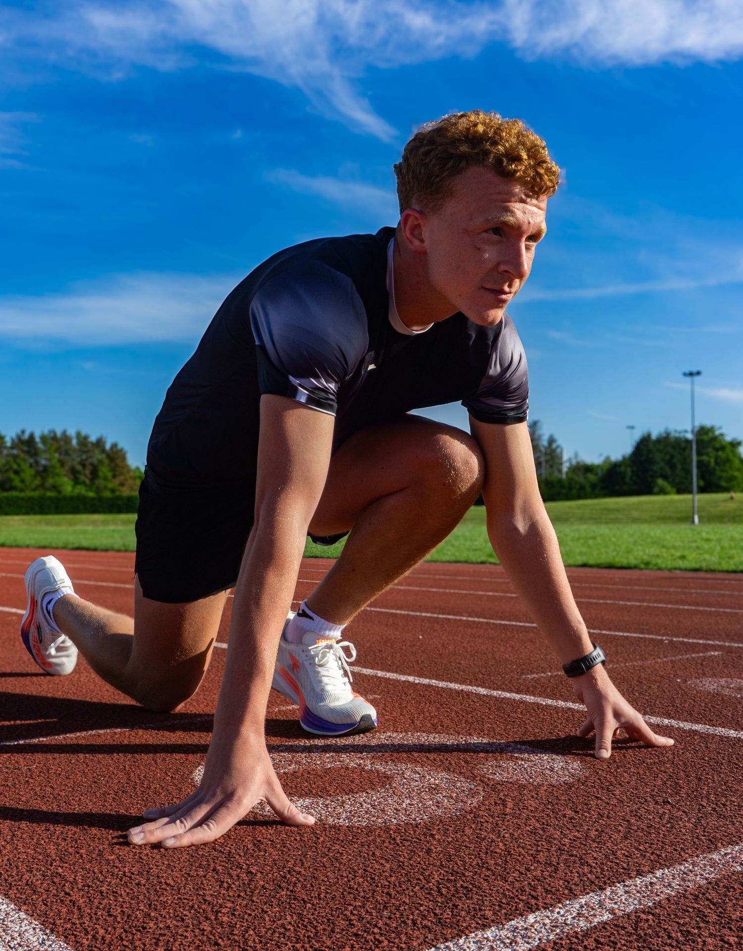 Young male athlete with curly hair in starting position on a red athletics track.
