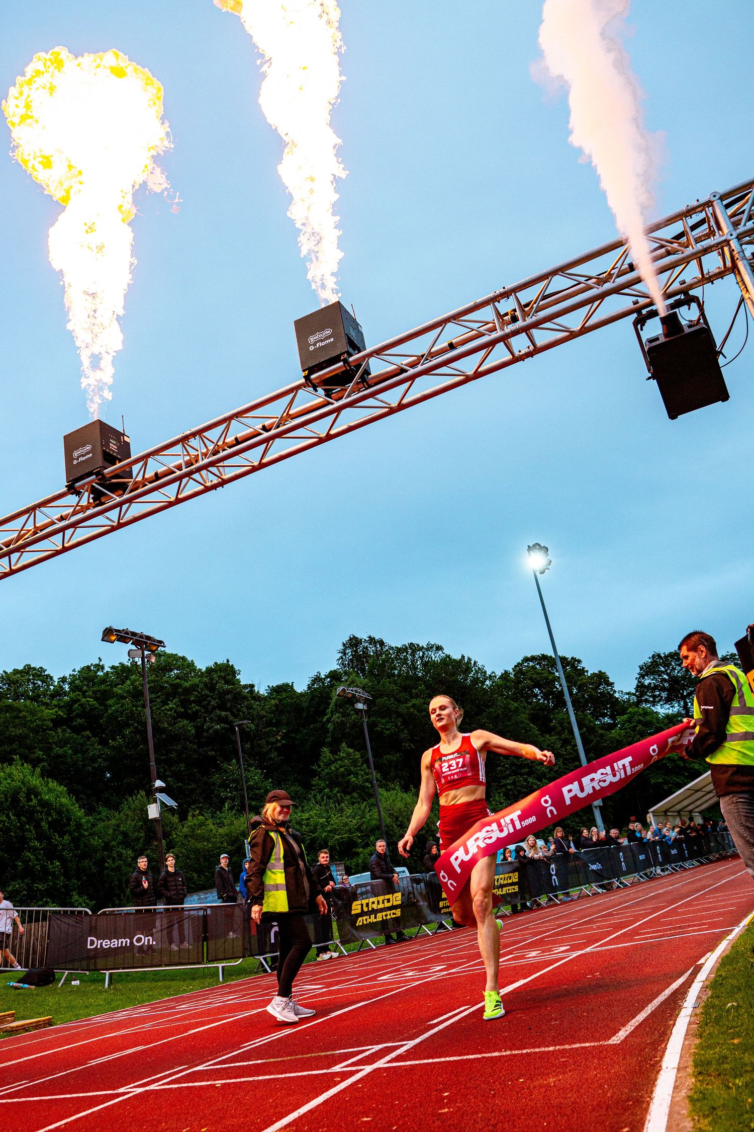 Female athlete breaks finish line tape at track event with dramatic pyrotechnic flames firing overhead at dusk.