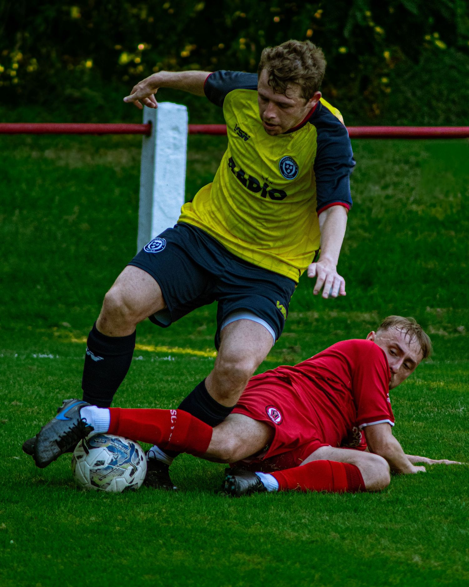Two soccer players battle for the ball during a match, one in yellow sliding tackle against a player in red kit.
