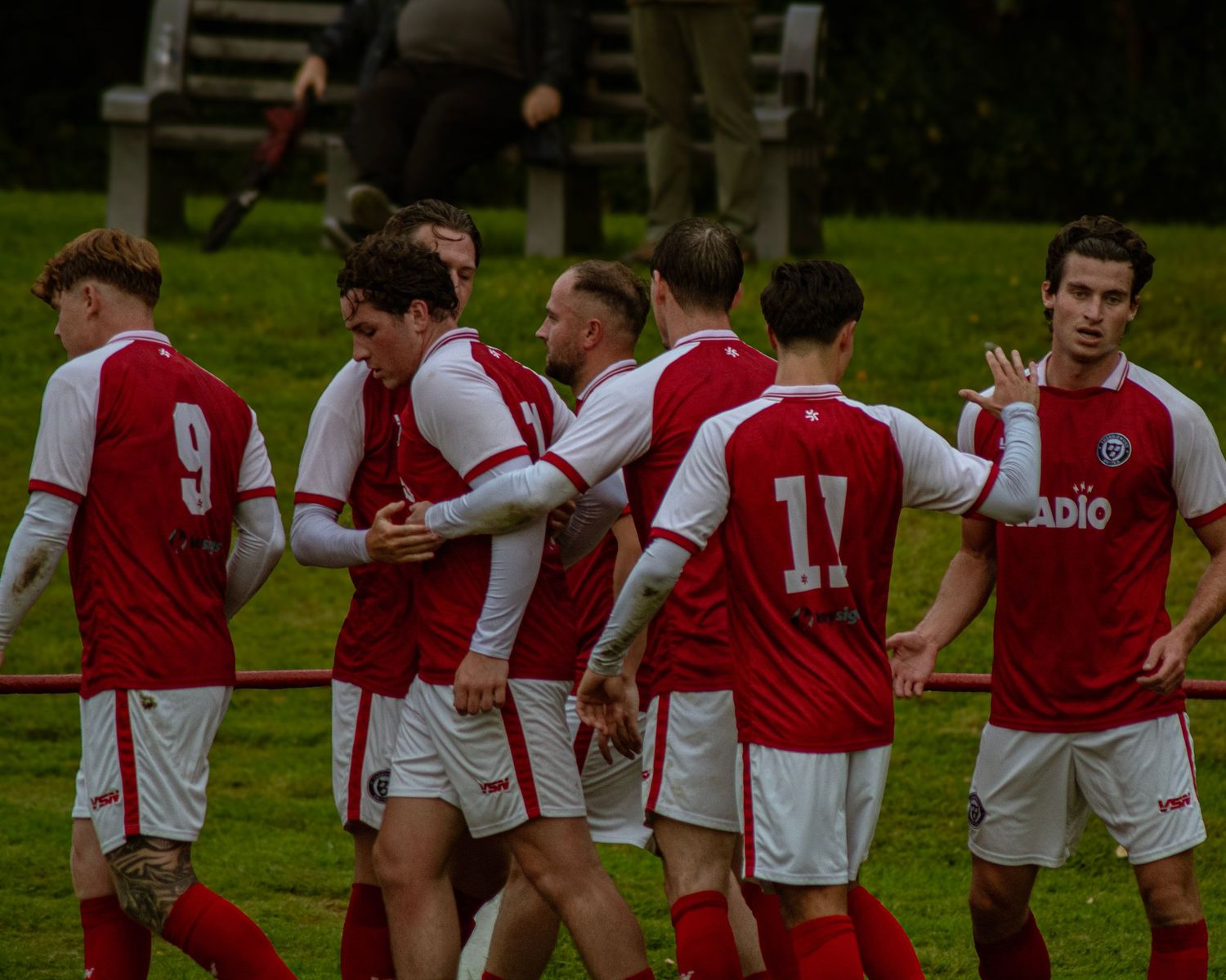 Red and white soccer players celebrate a goal together on a green grass pitch during a competitive match.