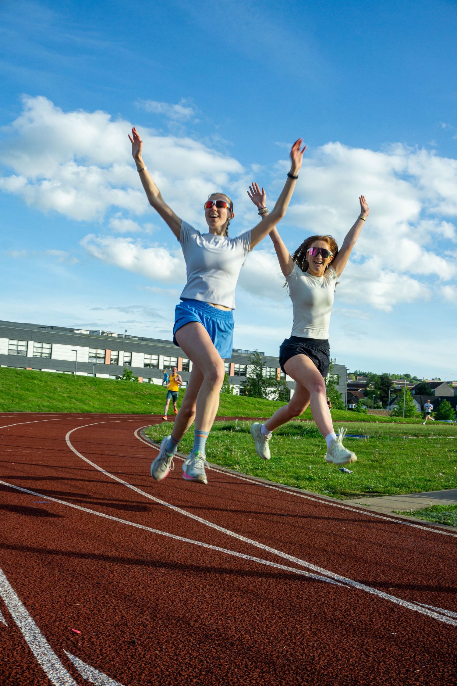 Two women joyfully jump with arms raised on a red running track under blue sky.