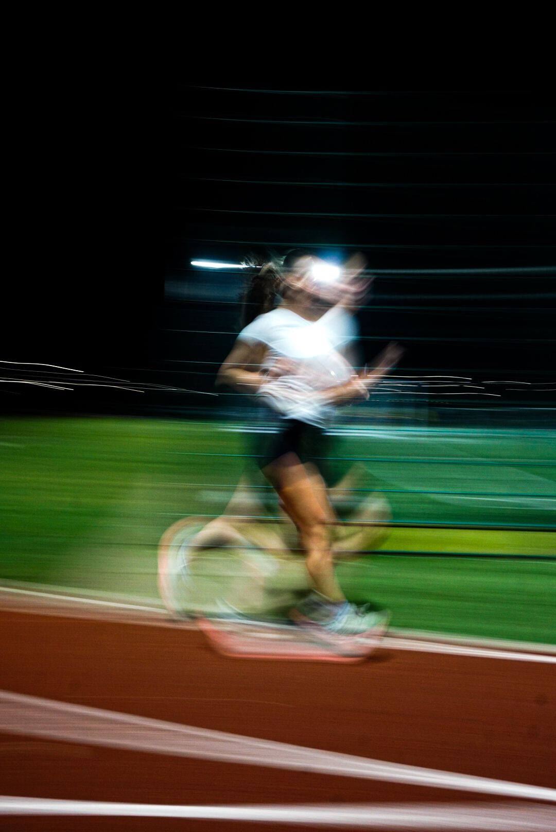 Blurred motion photograph of a female runner in white top sprinting on a red track at night.