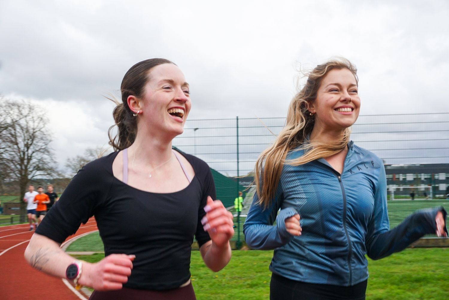 Two smiling women running together on an outdoor athletics track on a cloudy day.