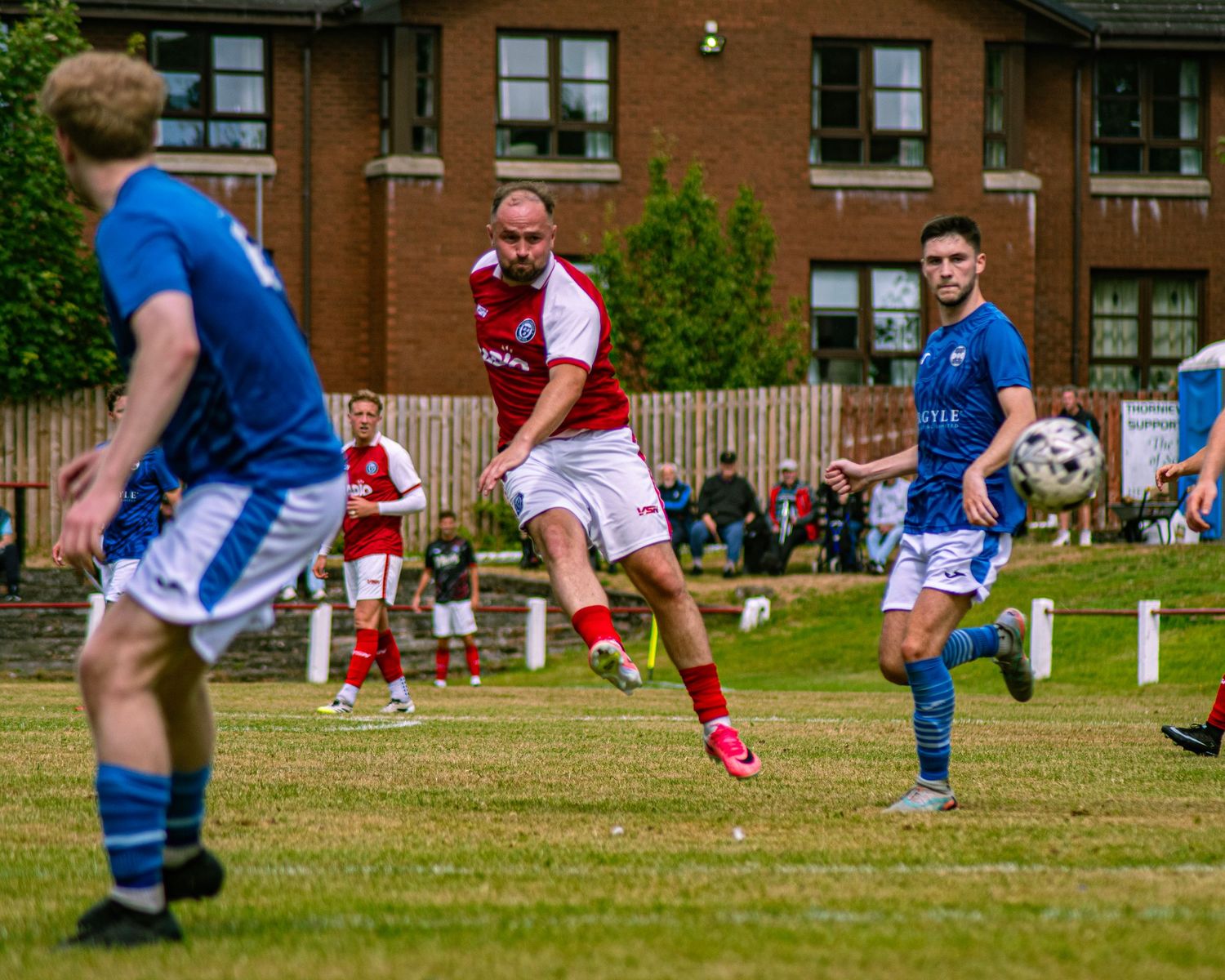 Soccer players in red and blue kits compete on a grass pitch with brick buildings in the background.