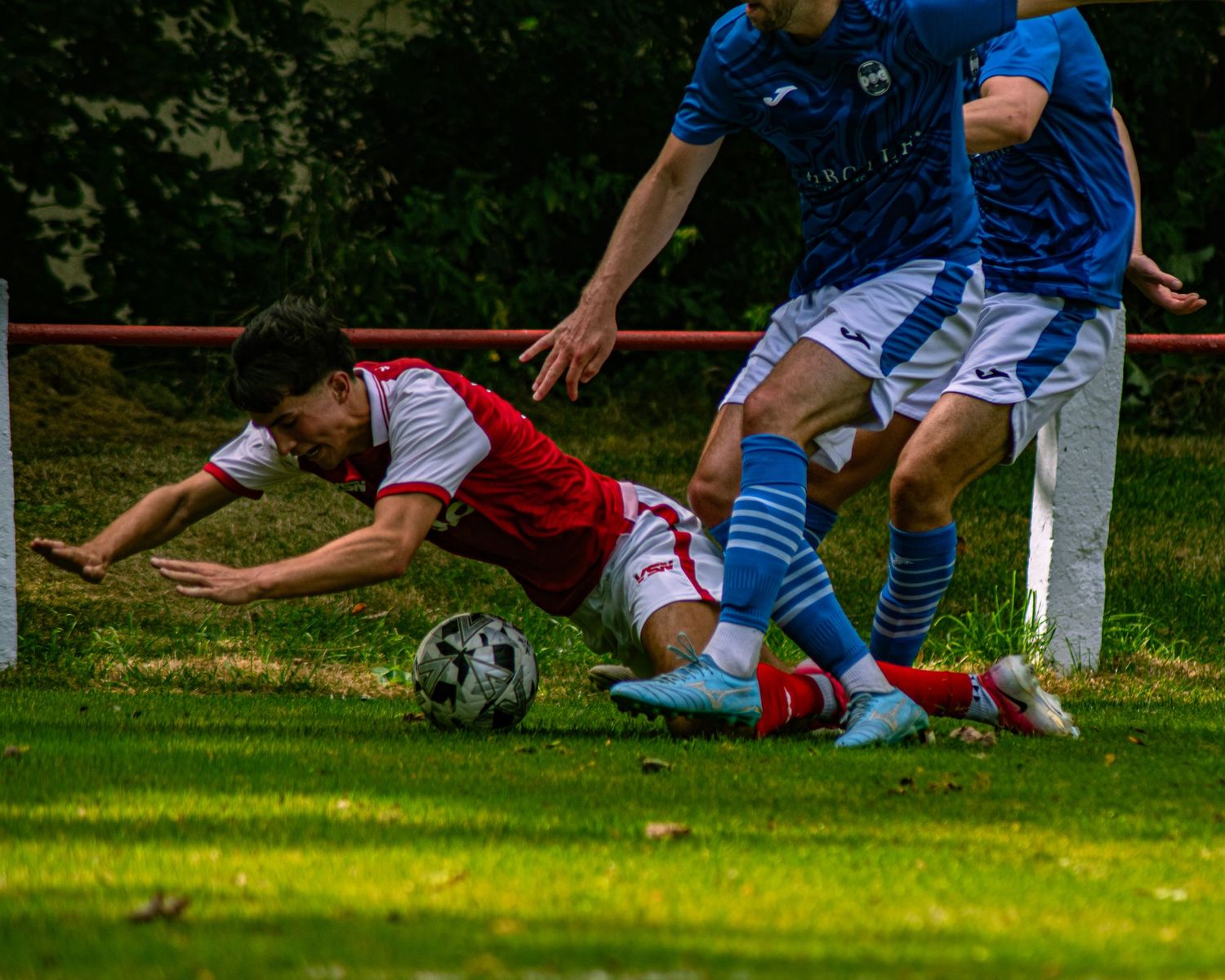 Football player in red kit diving for ball against blue-kitted opponents on grass pitch.