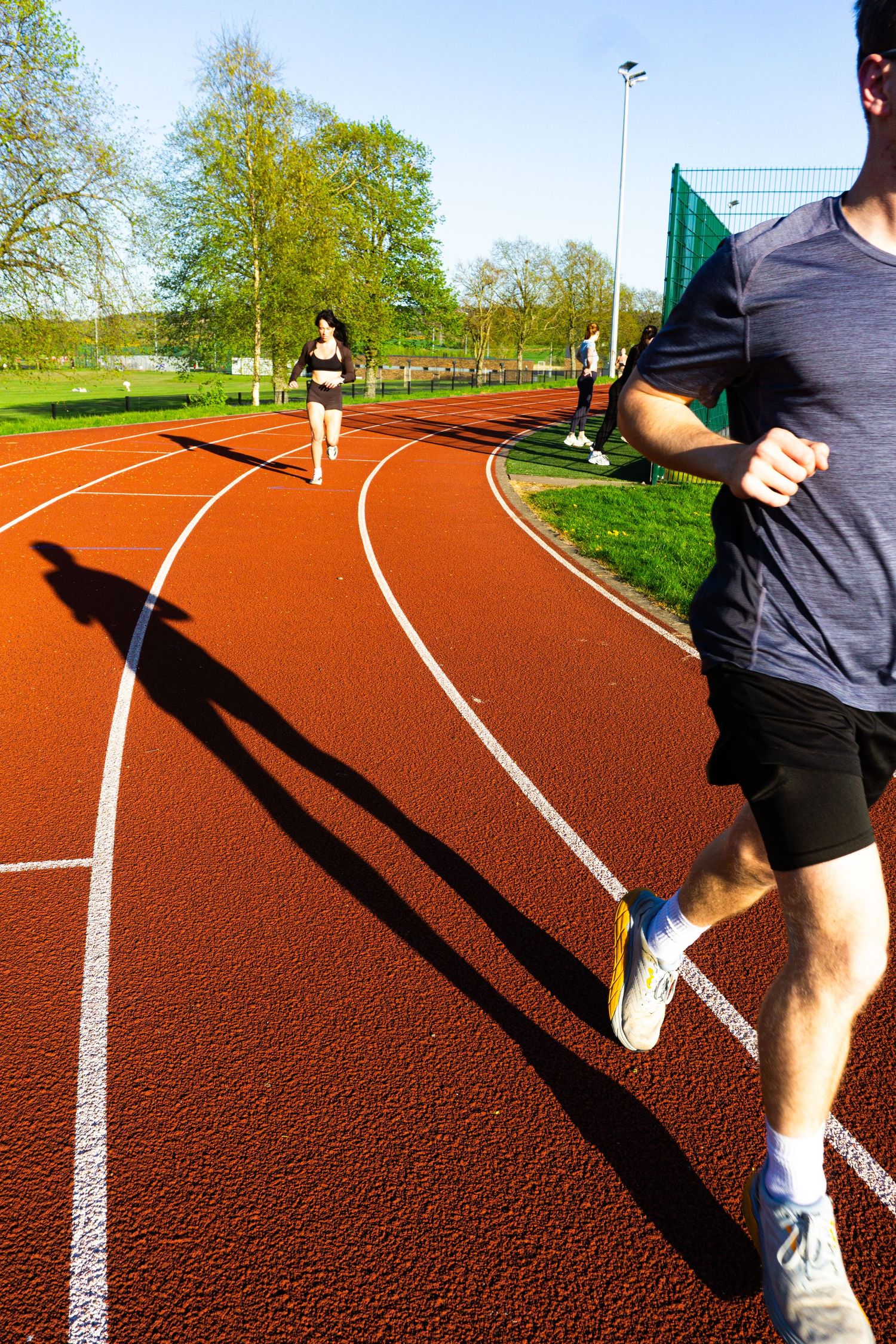 Runner casting long shadow on red athletics track with another runner in background.