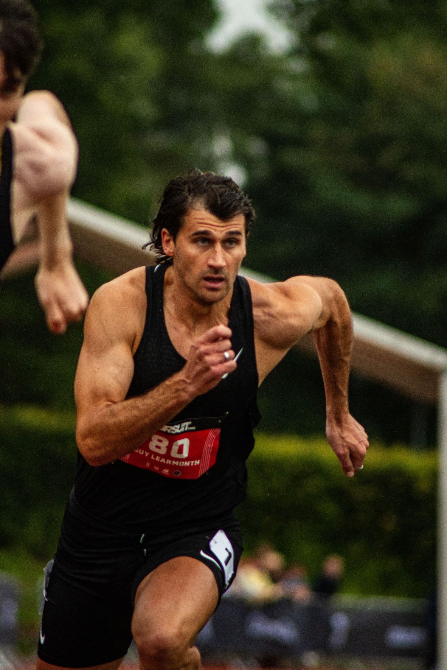 Male athlete wearing black tank top and bib number 80 sprinting intensely during a track and field competition.