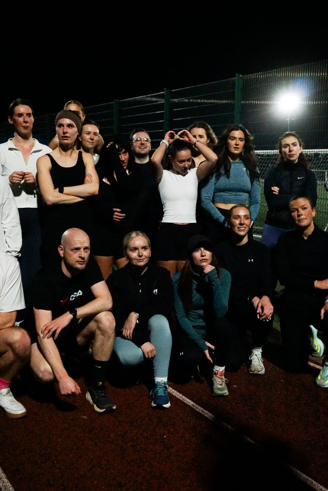 Group of diverse runners posing together on a floodlit outdoor running track at night.