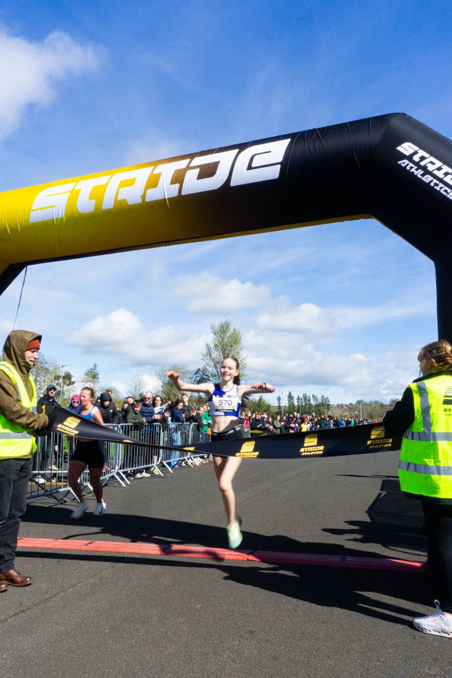 Female runner crosses the finish line under a Stride Athletics inflatable arch at an outdoor road race.