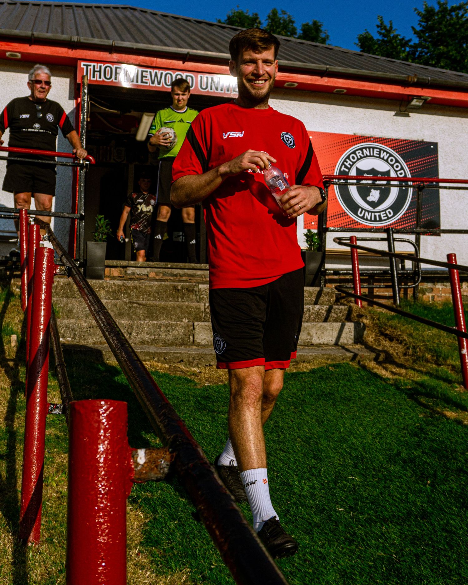 A smiling footballer in red kit walks out holding a water bottle at Thornlewood United FC ground.