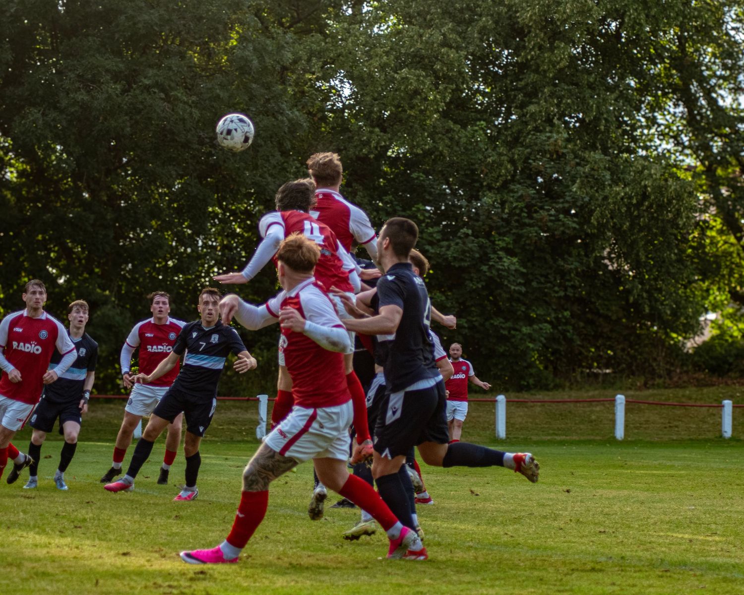 Soccer players compete for a header during an outdoor match, with red and black kits clashing near the goal area.