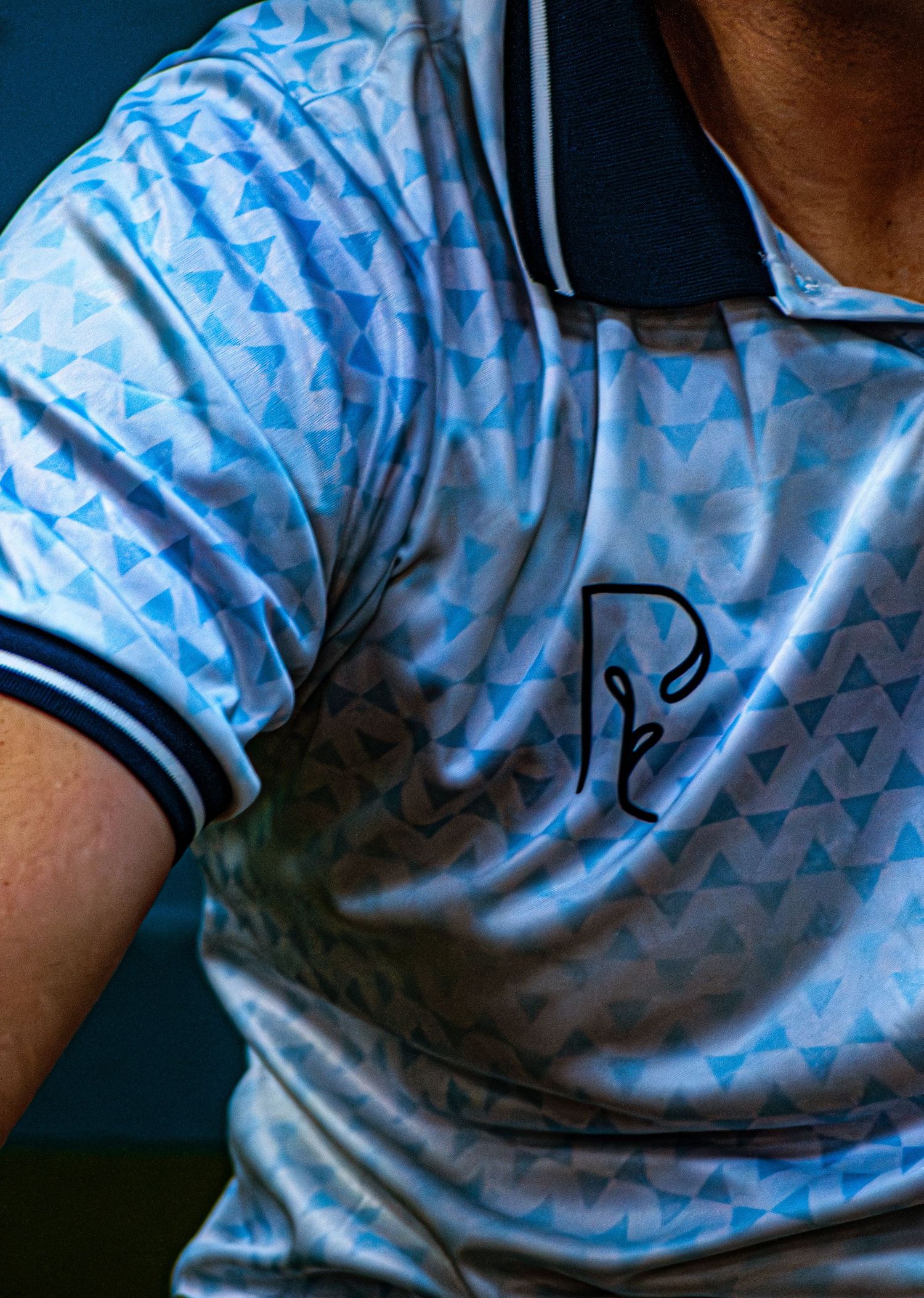 Blue geometric patterned polo shirt with navy collar and embroidered logo detail.