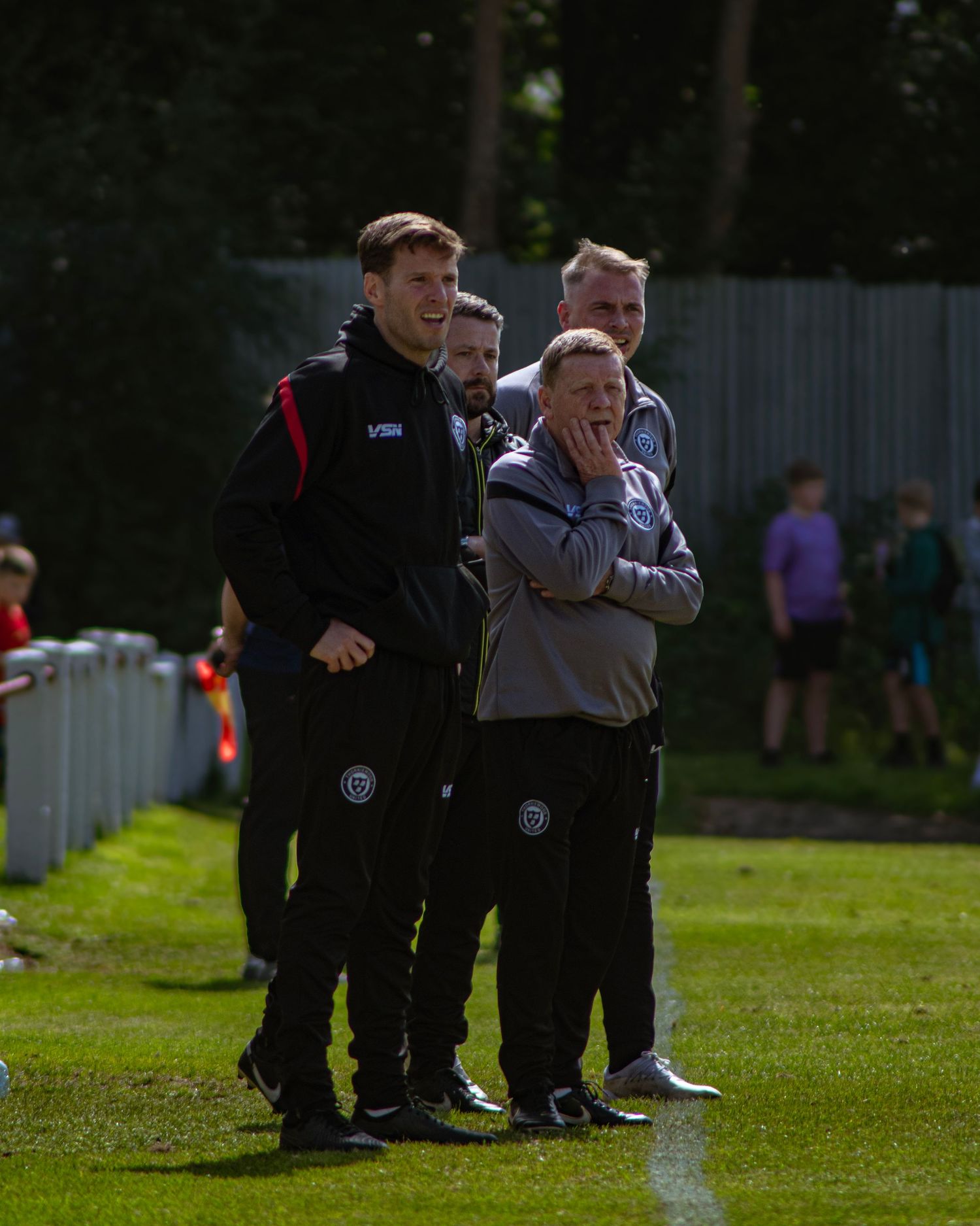 Football coaching staff in black tracksuits watch match action from the touchline on a sunny day.