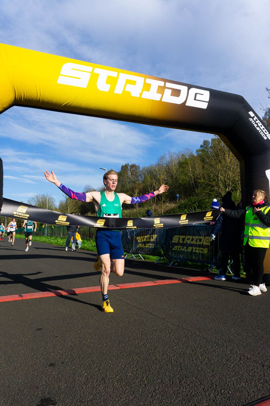 Male runner crosses finish line under a STRIDE Athletics inflatable arch with arms outstretched.