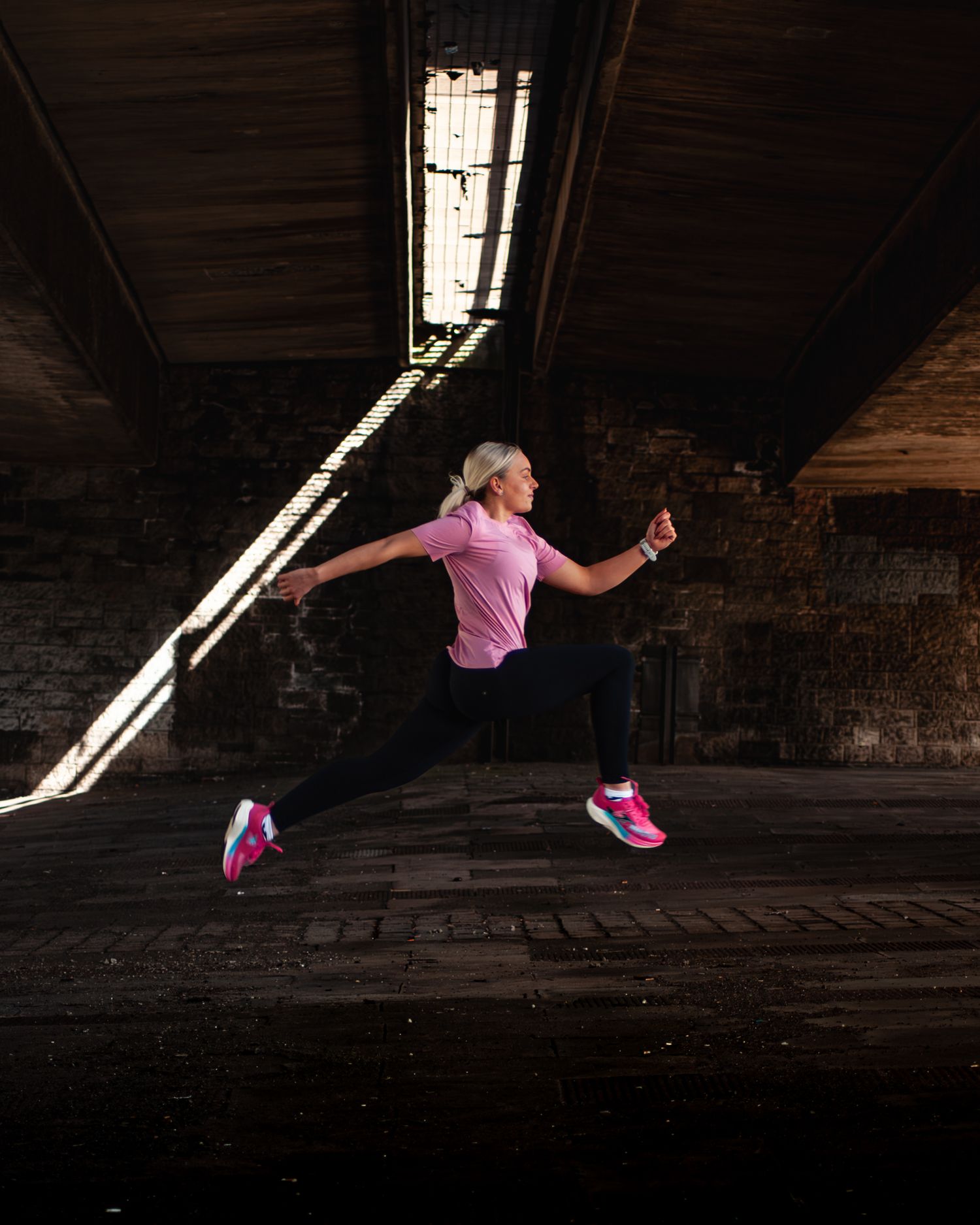 A blonde woman in a pink top and black leggings lunges dynamically in pink Joma sneakers inside a dark brick space.
