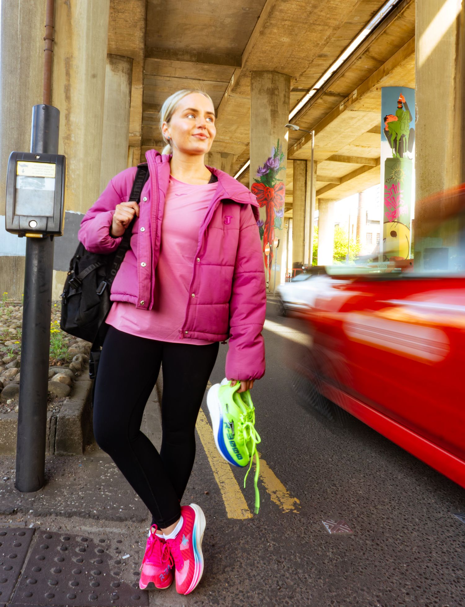 Blonde woman in pink puffer jacket carrying neon running shoes near busy urban underpass.