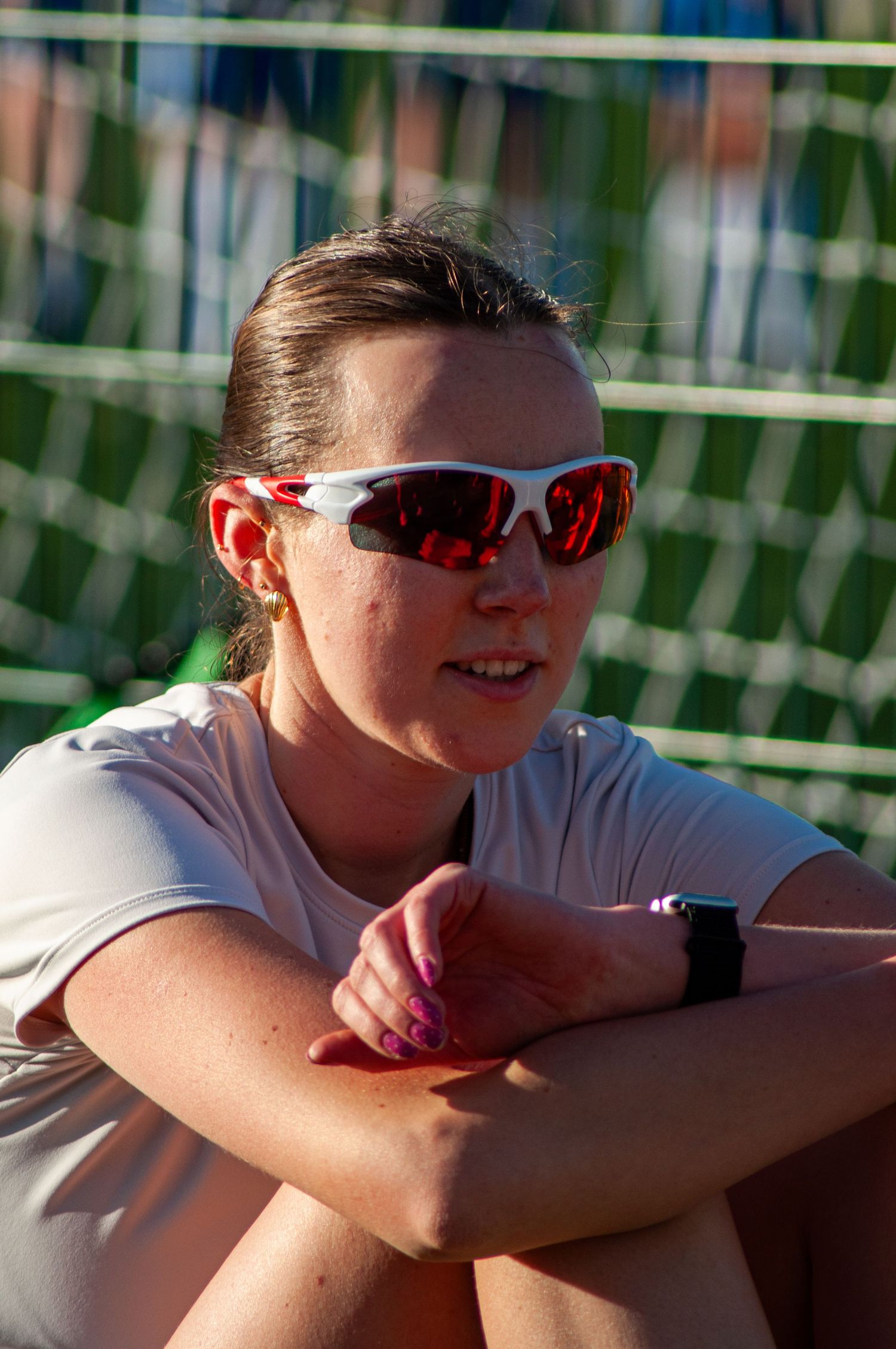 Female athlete resting with arms crossed wearing reflective red sports sunglasses near a green net.