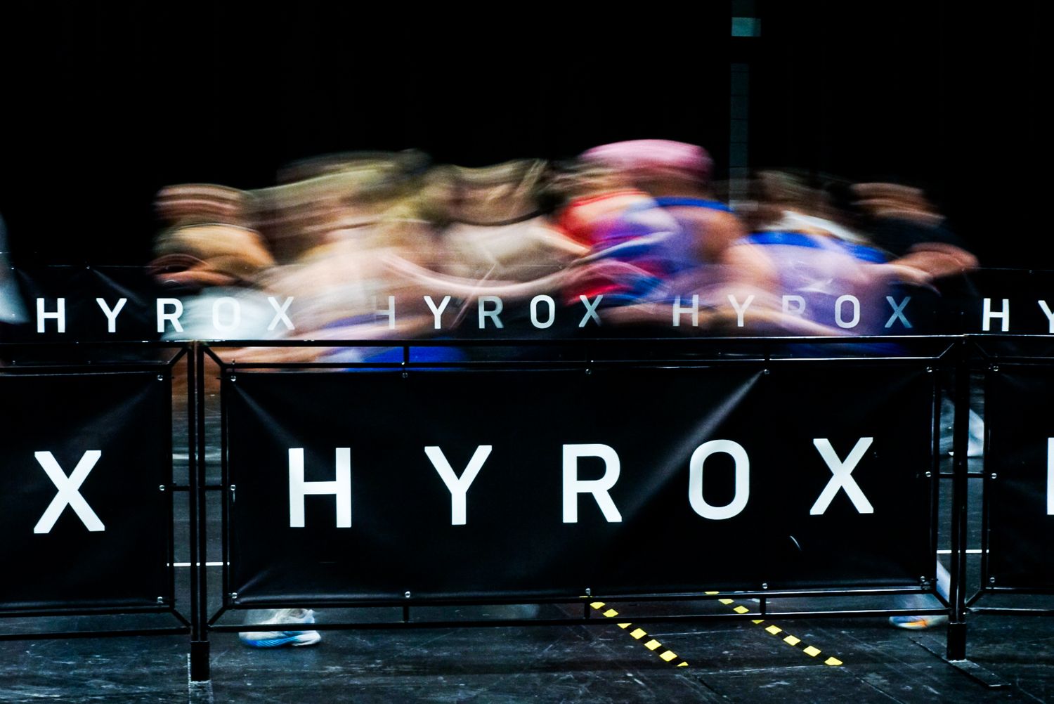 Motion-blurred athletes sprint past illuminated HYROX branded signage at an indoor competition.