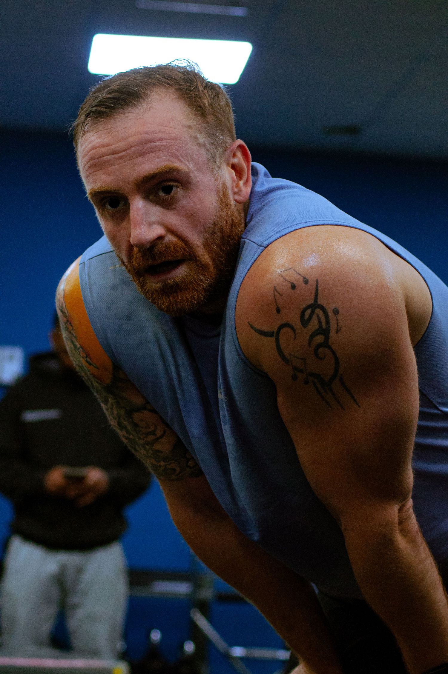 Muscular tattooed man with red beard resting between sets in gym.