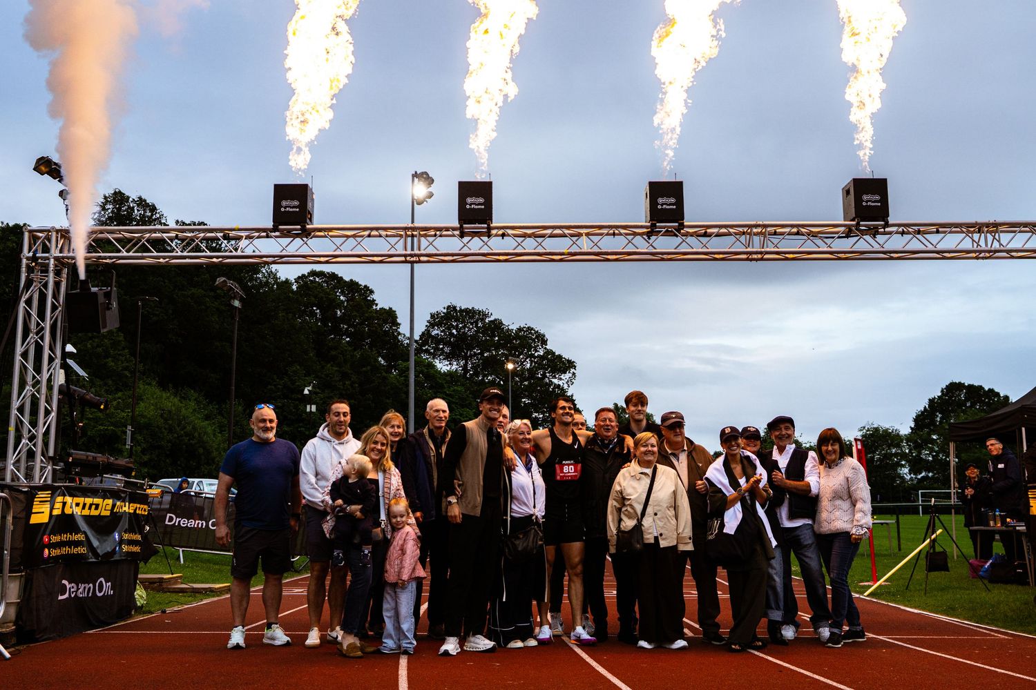 Group of people posing on a track under dramatic fire cannons at an outdoor athletics event.