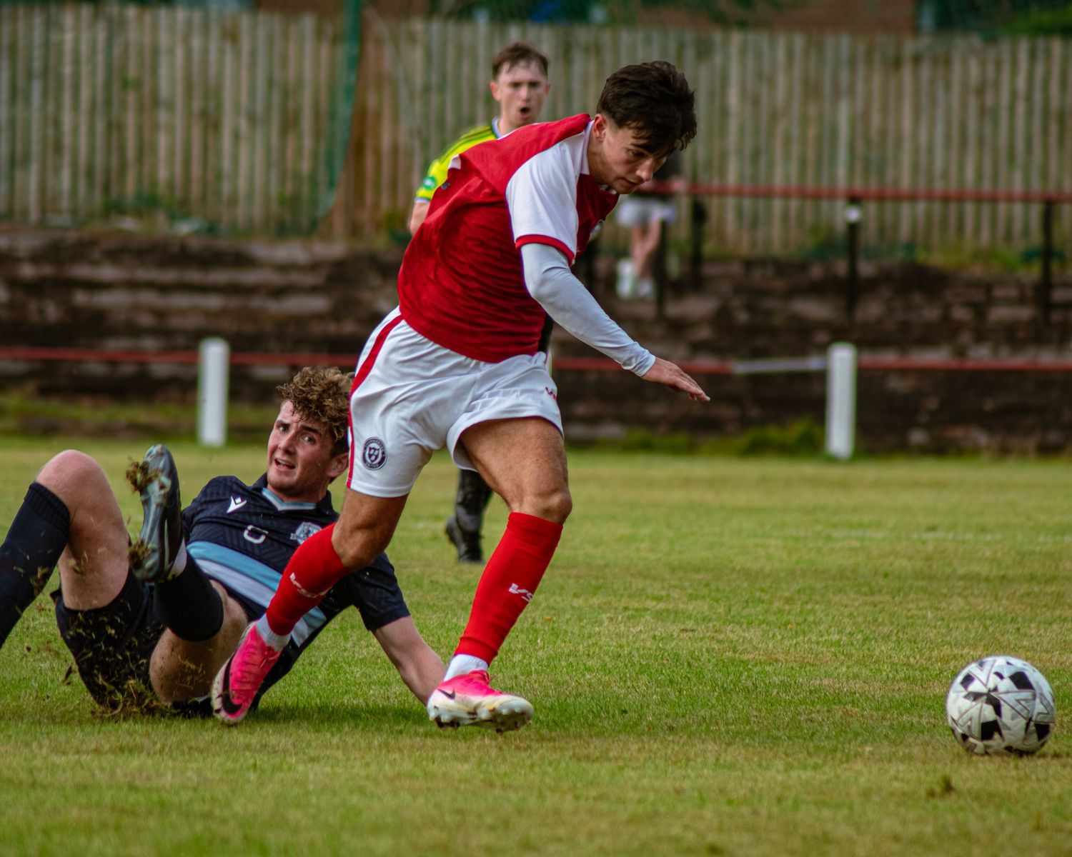Two soccer players compete for the ball during a match, one sliding on the grass while the other dribbles in a red kit.