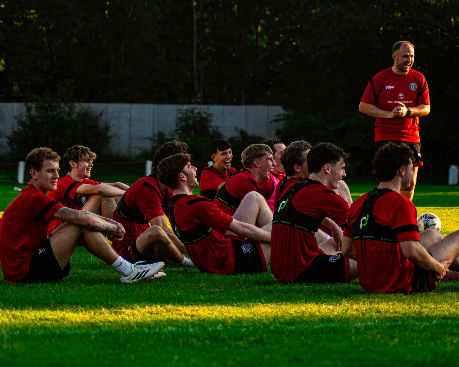 Soccer team in red jerseys sitting on grass listening to coach during outdoor training session in evening light.