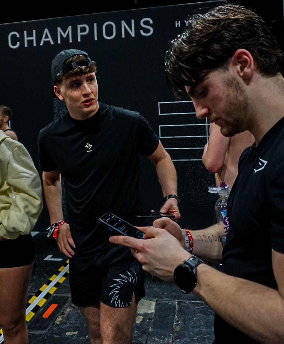 Two male athletes in black sportswear discuss results at a HYROX Champions event.