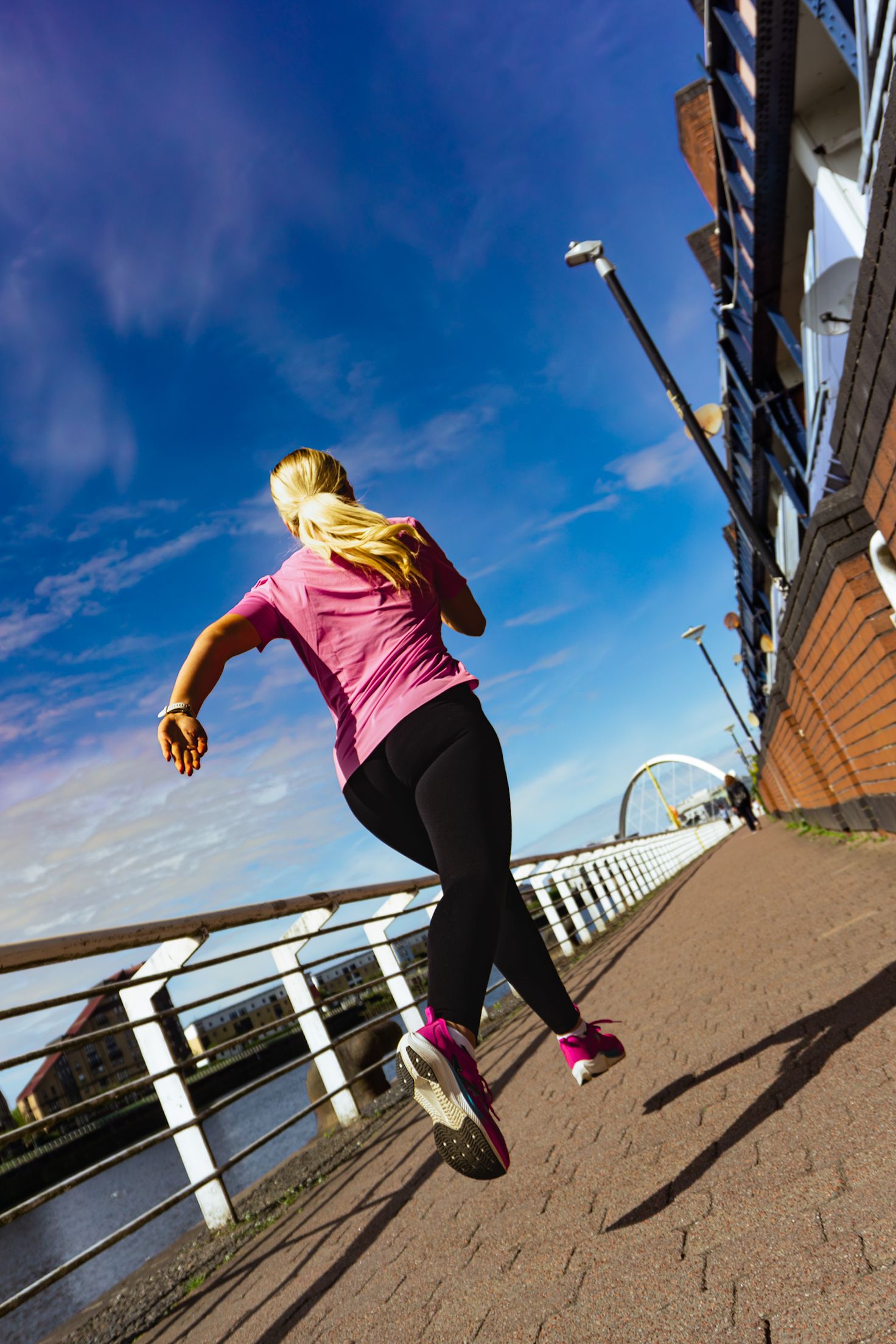 Woman in pink shirt and black leggings running along urban waterfront path under blue sky.
