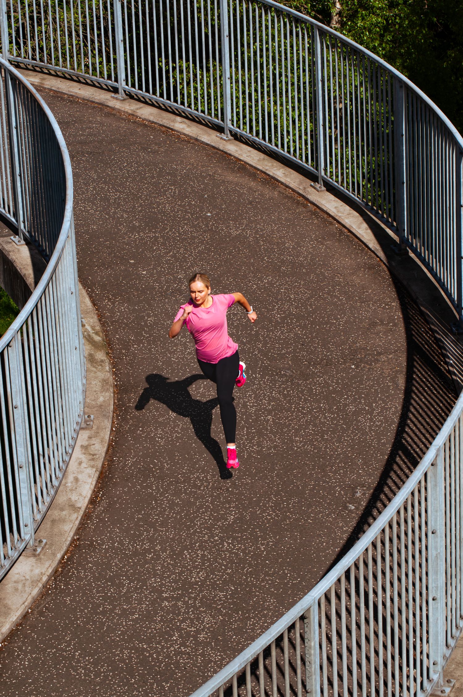 An aerial view of a female runner in a pink top and Joma sneakers jogging along a curved spiral bridge path.