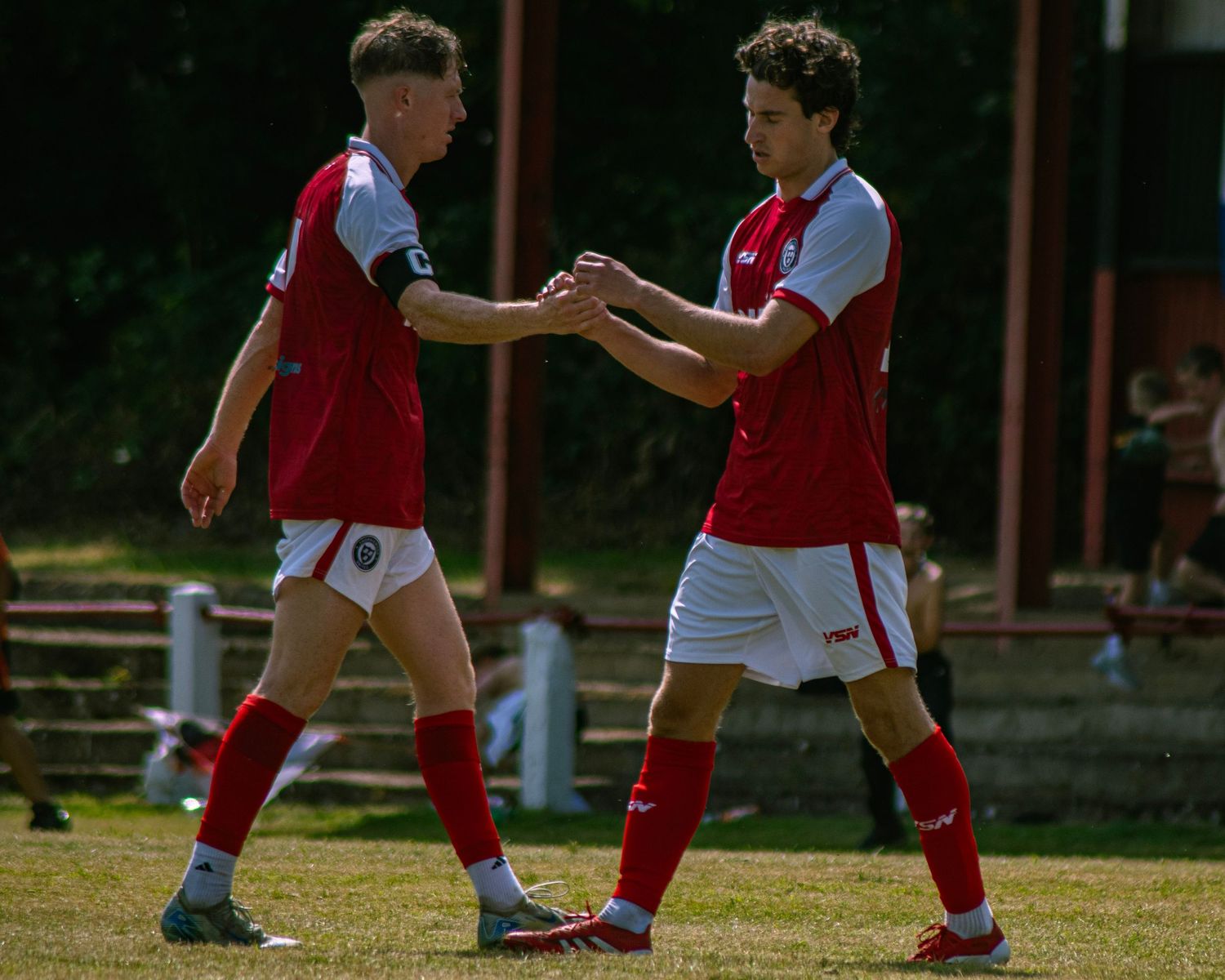 Two youth soccer players in red and white uniforms shake hands on a sunny green field during a match.