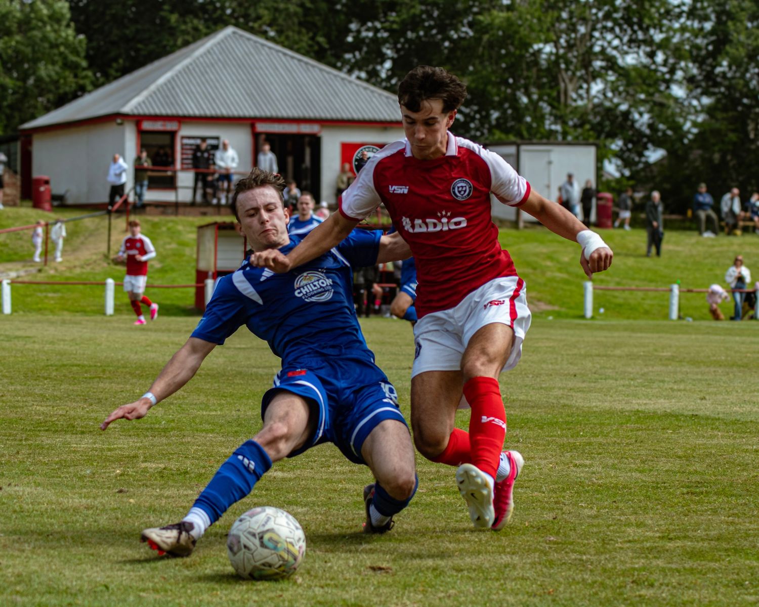A player in red and white tackles a blue-kitted opponent during a competitive grassroots football match.