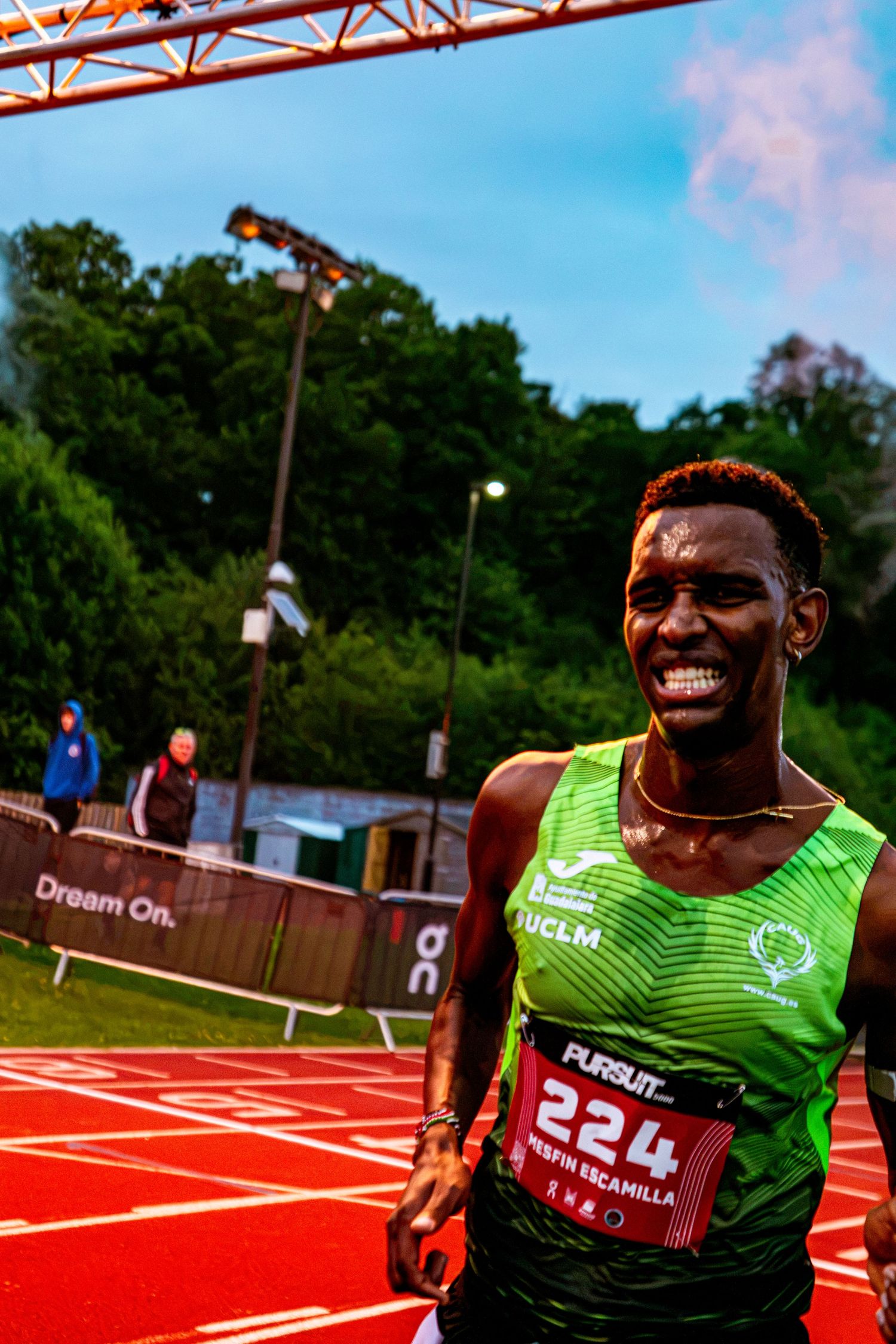 Smiling male runner wearing green UCLM jersey with bib number 224 celebrates finishing a track race at outdoor athletics event.
