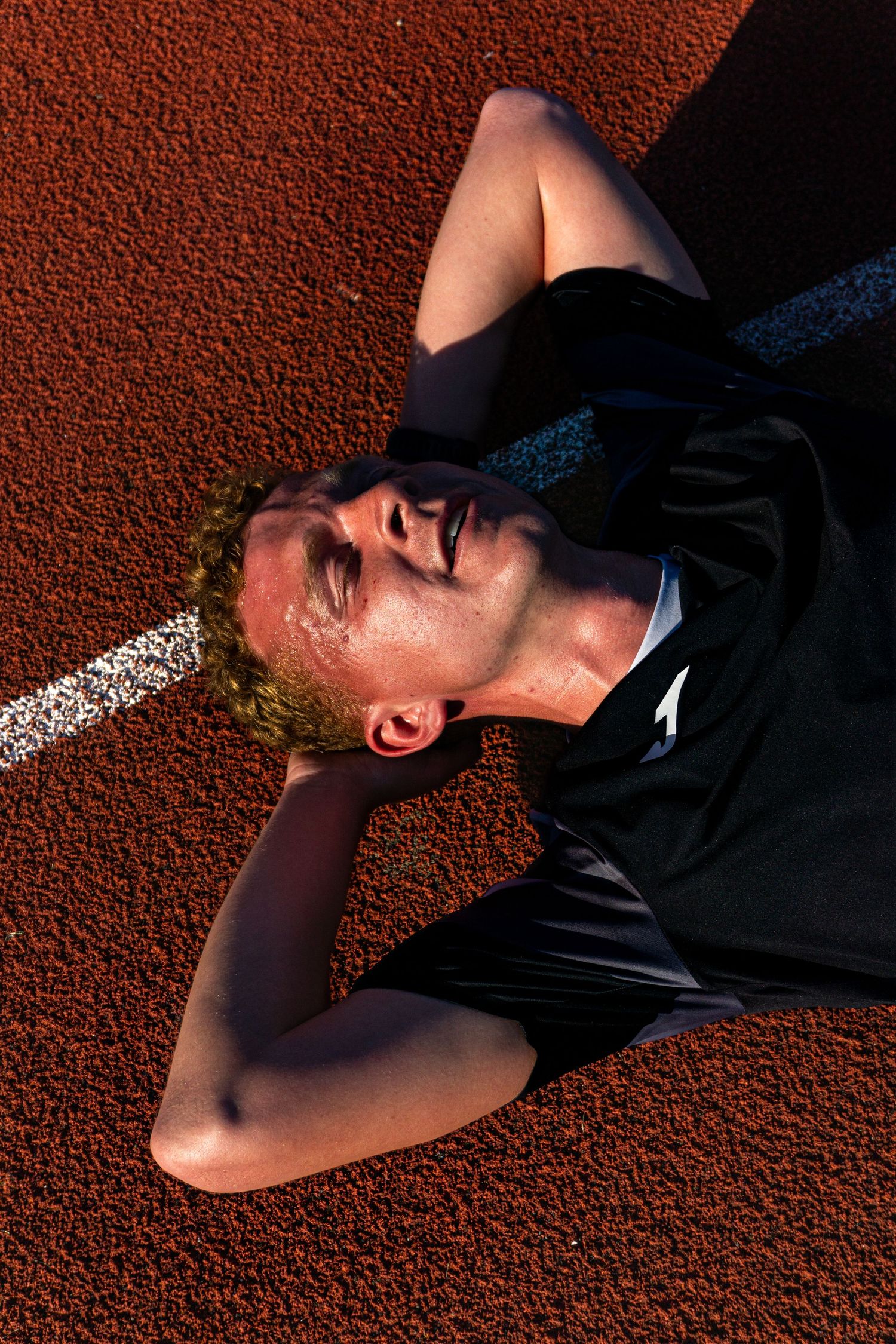 Exhausted male athlete lying on a red running track with hands behind head, eyes closed in sunlight.