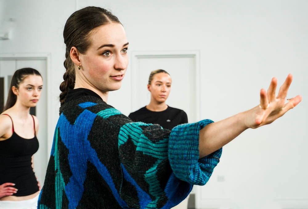 A young female dancer in a colorful blue jacket extends her arm gracefully during a dance rehearsal in a bright studio.