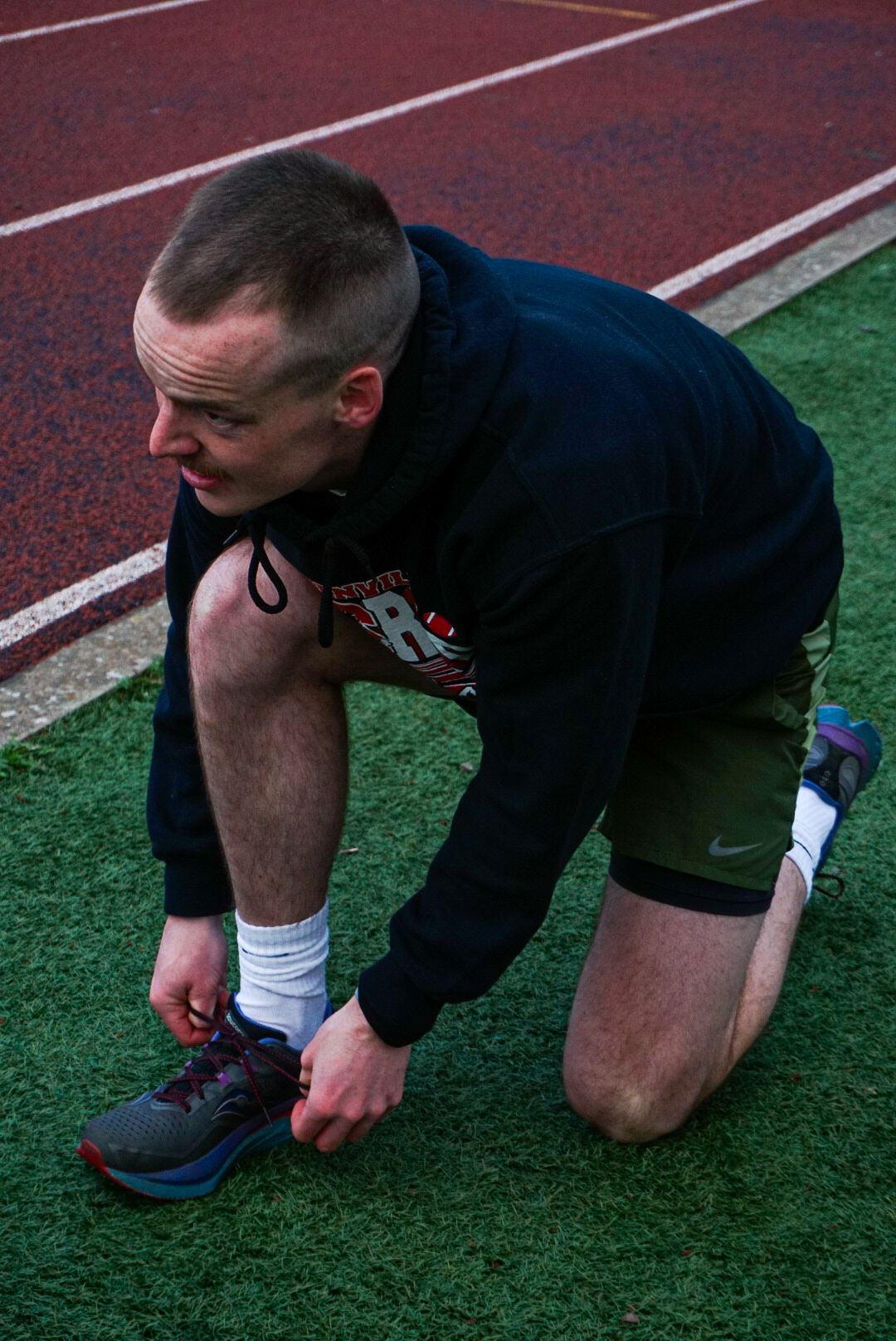 Male athlete kneeling on grass beside red track tying his running shoes.