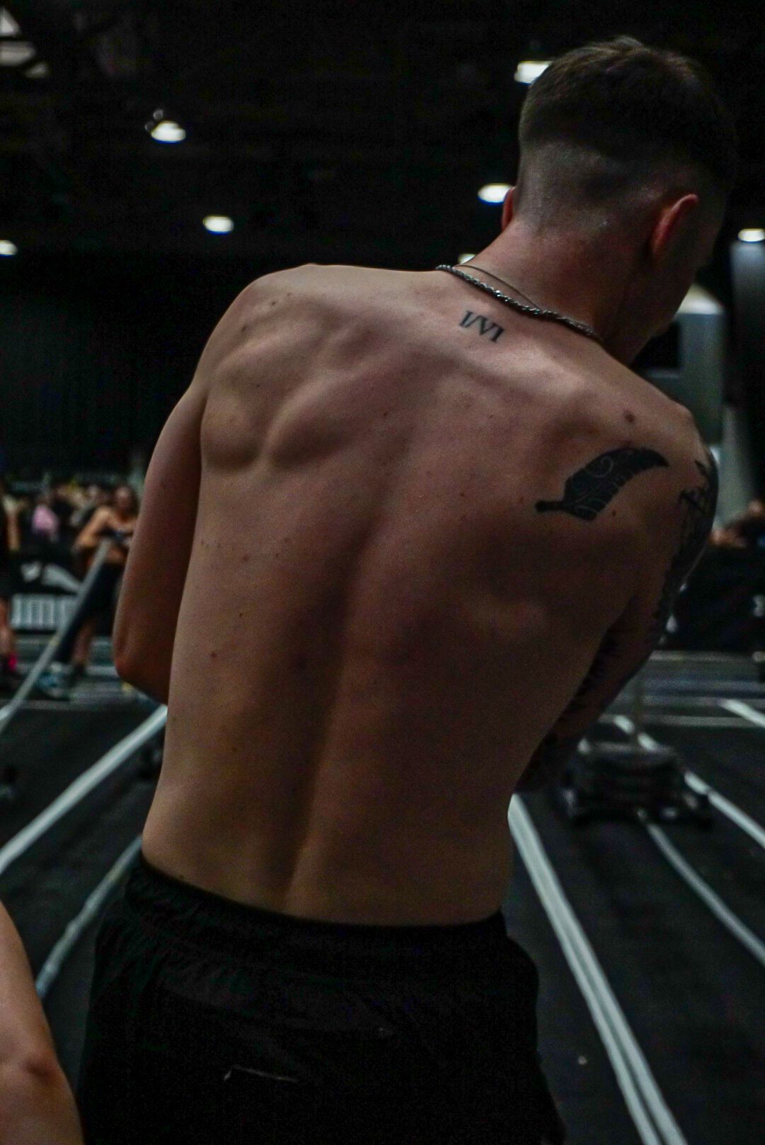 Muscular shirtless man with back tattoos standing in a gym during a CrossFit competition.
