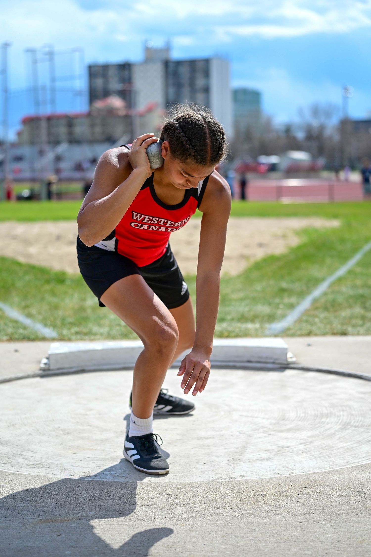 Female athlete in Western Canada jersey preparing to throw shot put at outdoor track competition.