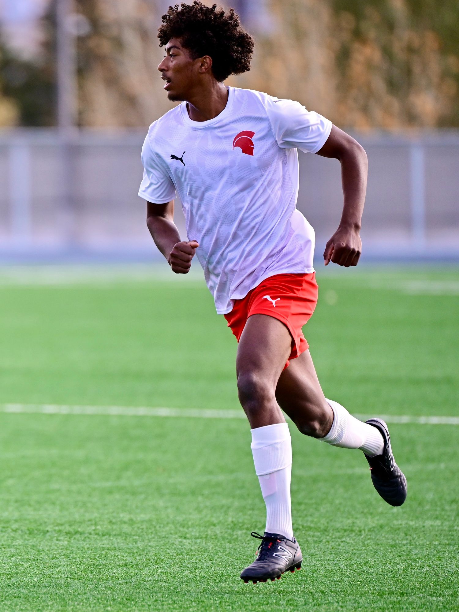 Young soccer player in white and red Spartan uniform sprinting on green turf field during game.